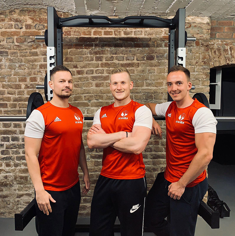Drei Fitness Trainer in Roten oberteilen und schwarzen Hosen stehen vor einer Hantelbank